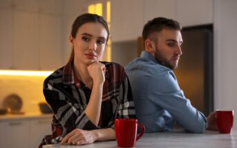 A woman and a man sit back-to-back at a kitchen counter, both appearing thoughtful and distant. Two red mugs are on the counter in front of them. The kitchen is softly lit, with warm tones and modern decor.