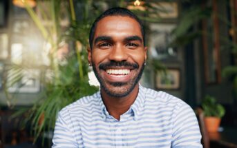 A young man with short hair and a beard smiles brightly while wearing a blue and white striped shirt, sitting indoors with green plants and soft lighting in the background.