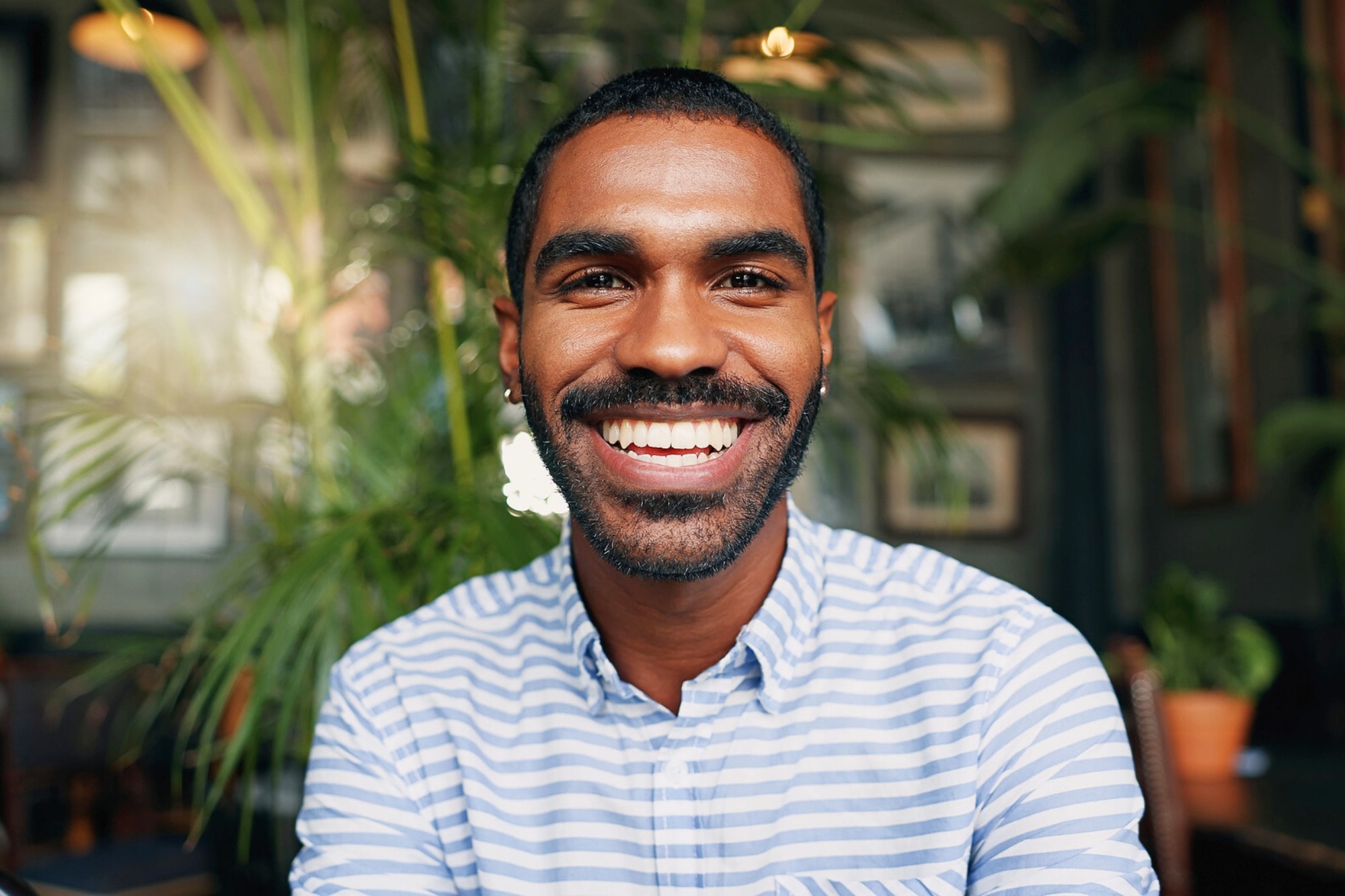 A young man with short hair and a beard smiles brightly while wearing a blue and white striped shirt, sitting indoors with green plants and soft lighting in the background.