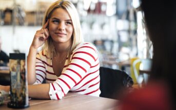 A woman with blonde hair wearing a white and red striped top sits at a table in a cafe, smiling and resting her head on her hand while listening to someone across from her.