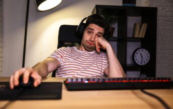 A young man wearing headphones and a striped shirt sits at a desk with a keyboard and mouse, looking bored and leaning his head on his hand, with a clock and shelves in the background.