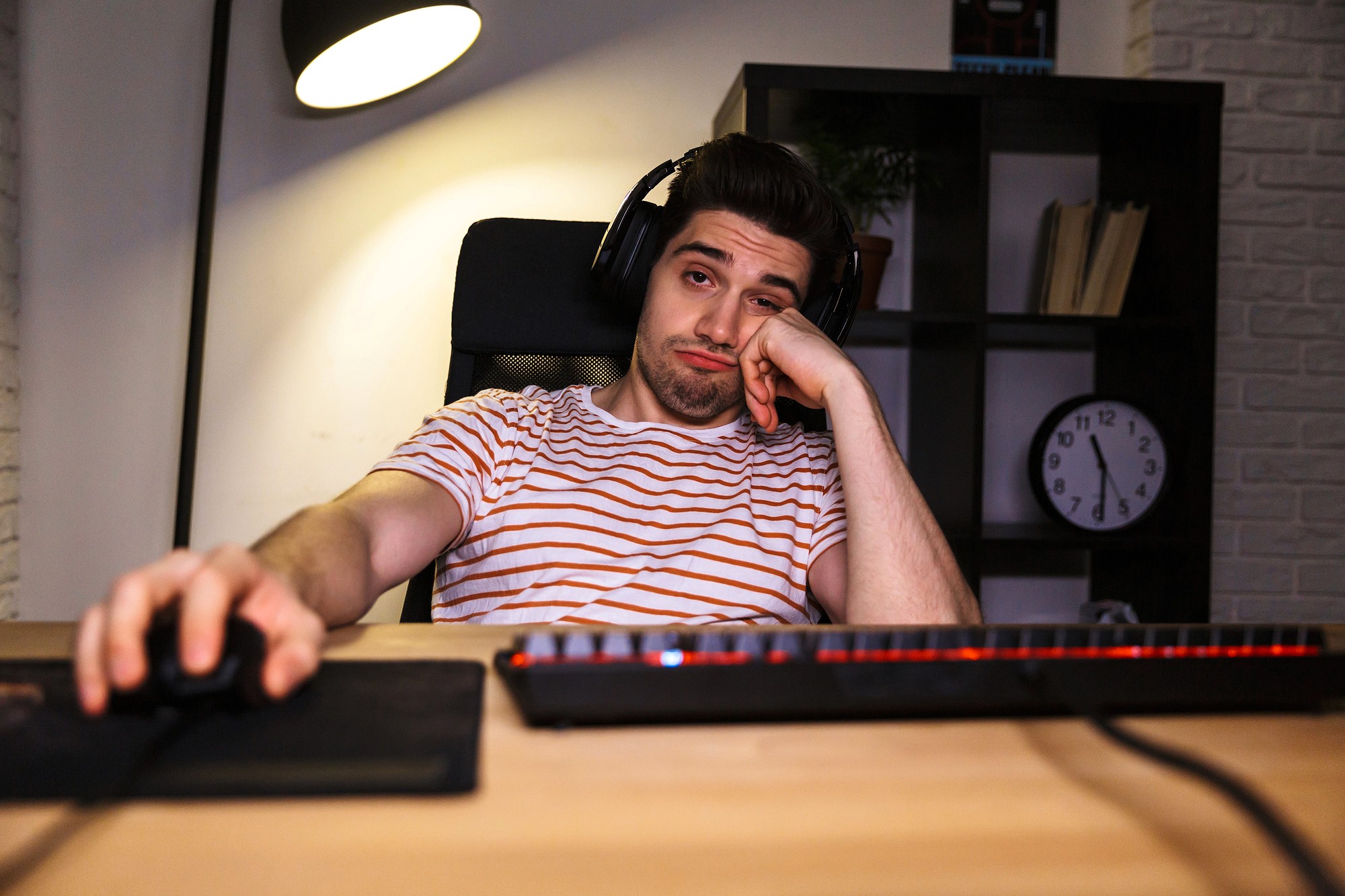 A young man wearing headphones and a striped shirt sits at a desk with a keyboard and mouse, looking bored and leaning his head on his hand, with a clock and shelves in the background.