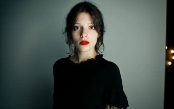 A young woman with pale skin, dark hair, and red lipstick stands against a plain, gray background, wearing a black top with lace details and gazing directly at the camera with a neutral expression.
