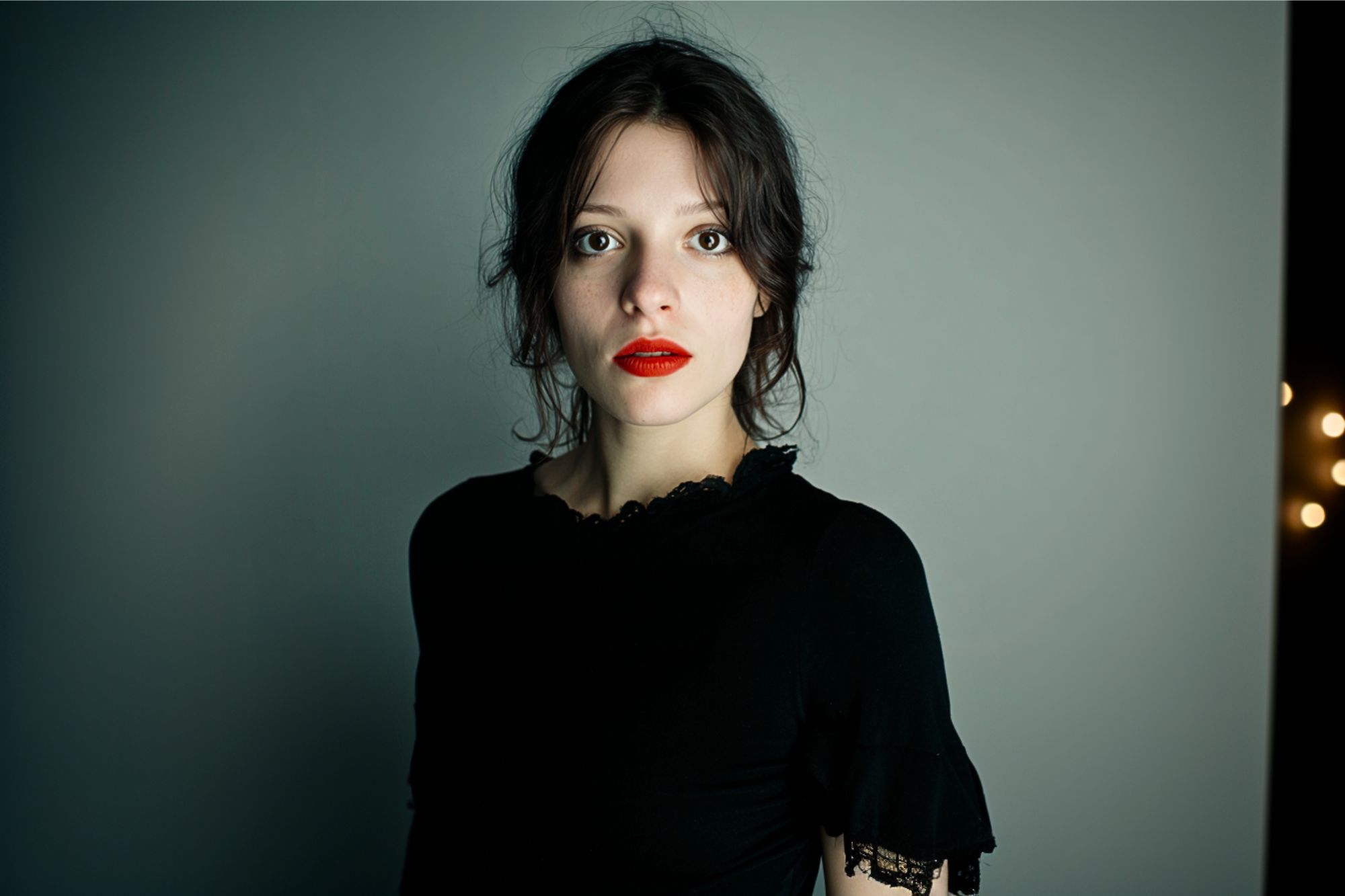 A young woman with pale skin, dark hair, and red lipstick stands against a plain, gray background, wearing a black top with lace details and gazing directly at the camera with a neutral expression.
