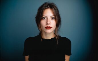 A young woman with long brown hair, red lipstick, and a black top stands against a gradient dark blue background, looking directly at the camera with a neutral expression.