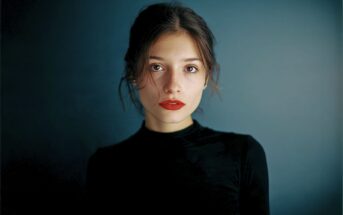 A young woman with light skin, red lipstick, and dark hair pulled back, wearing a black turtleneck, stands against a dark, softly lit blue background, gazing directly at the camera with a neutral expression.