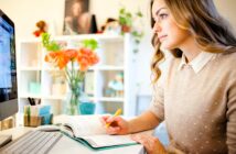 A woman with long hair sits at a desk, writing in a notebook while looking at her computer screen. She holds a yellow pen. In the background, there's a vase of orange flowers and bookshelves filled with various items.