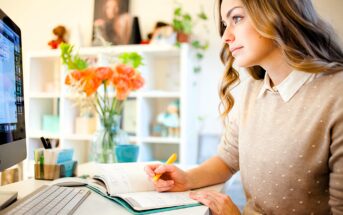 A woman with long hair sits at a desk, writing in a notebook while looking at her computer screen. She holds a yellow pen. In the background, there's a vase of orange flowers and bookshelves filled with various items.