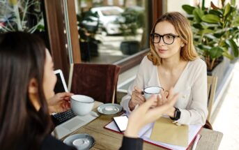 Two women sit at an outdoor café table, having a conversation over coffee. One woman faces the camera, wearing glasses, while the other is seen from behind. Notebooks, a laptop, and a phone are on the table.