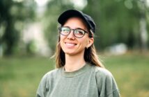 A young woman wearing glasses, a black cap, and an olive green sweatshirt smiles outdoors with a blurred green background.