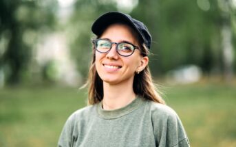 A young woman wearing glasses, a black cap, and an olive green sweatshirt smiles outdoors with a blurred green background.