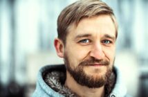 A man with light brown hair, a beard, and blue eyes wears a gray hooded jacket with a furry lining, smiling softly against a blurred outdoor background.