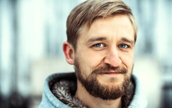 A man with light brown hair, a beard, and blue eyes wears a gray hooded jacket with a furry lining, smiling softly against a blurred outdoor background.