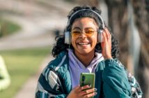 A smiling person with curly hair, wearing yellow-tinted glasses, large headphones, and a green jacket, holds a smartphone while standing outdoors on a sunny day.
