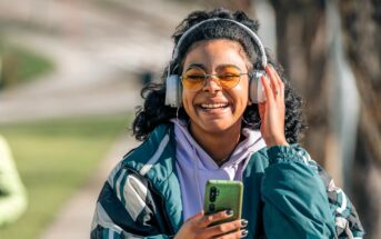 A smiling person with curly hair, wearing yellow-tinted glasses, large headphones, and a green jacket, holds a smartphone while standing outdoors on a sunny day.