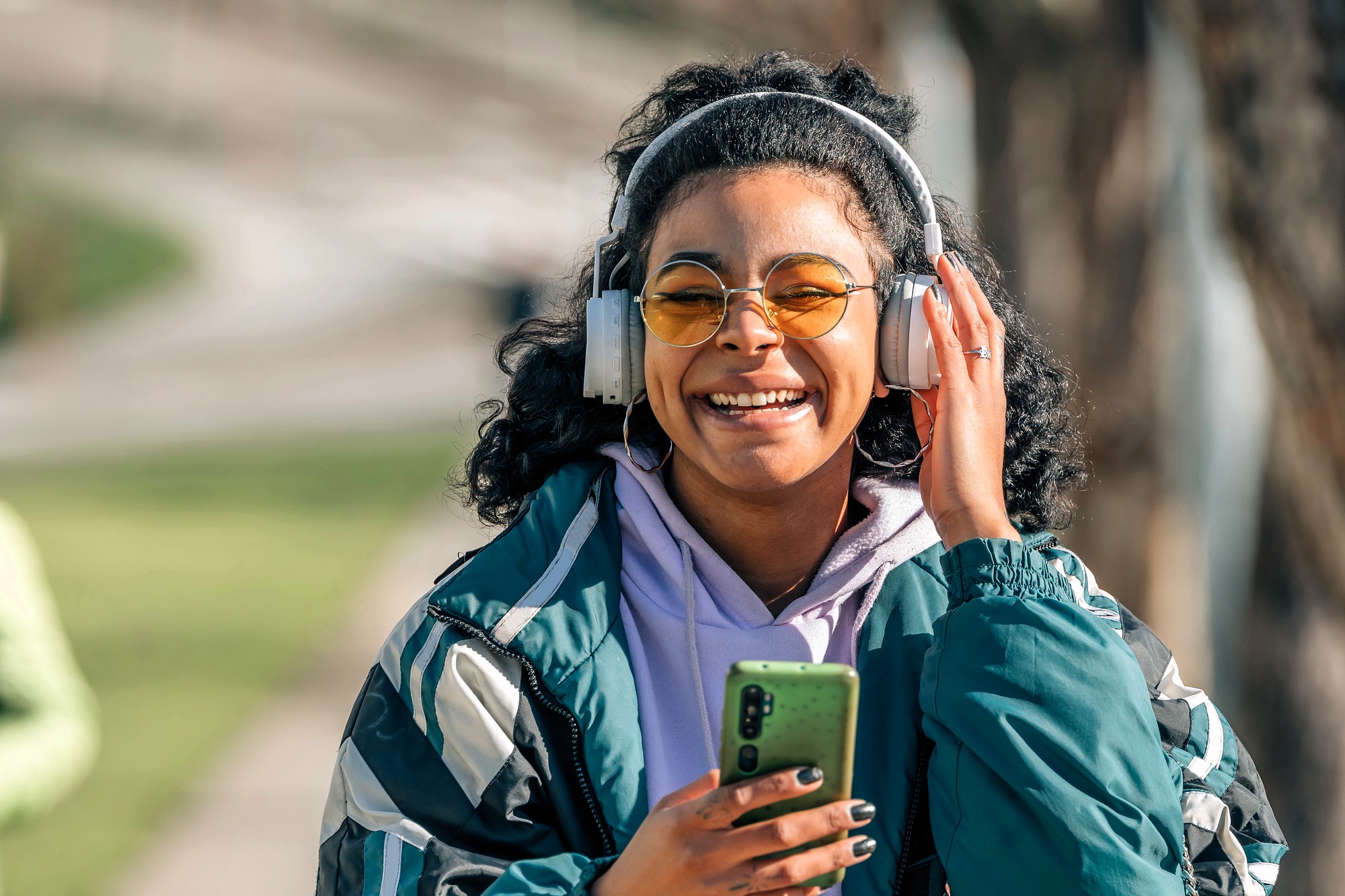 A smiling person with curly hair, wearing yellow-tinted glasses, large headphones, and a green jacket, holds a smartphone while standing outdoors on a sunny day.