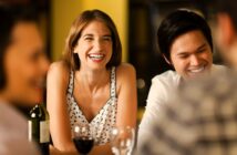 A woman and a man sit at a table, laughing and smiling, with glasses of wine in front of them. Two other people are blurred in the foreground, and the background is softly lit and out of focus.