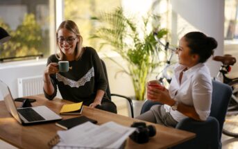 Two women sit at a desk in an office, smiling and chatting while holding mugs. Laptops, notebooks, and documents are on the desk, and a bicycle and plant are visible in the background.