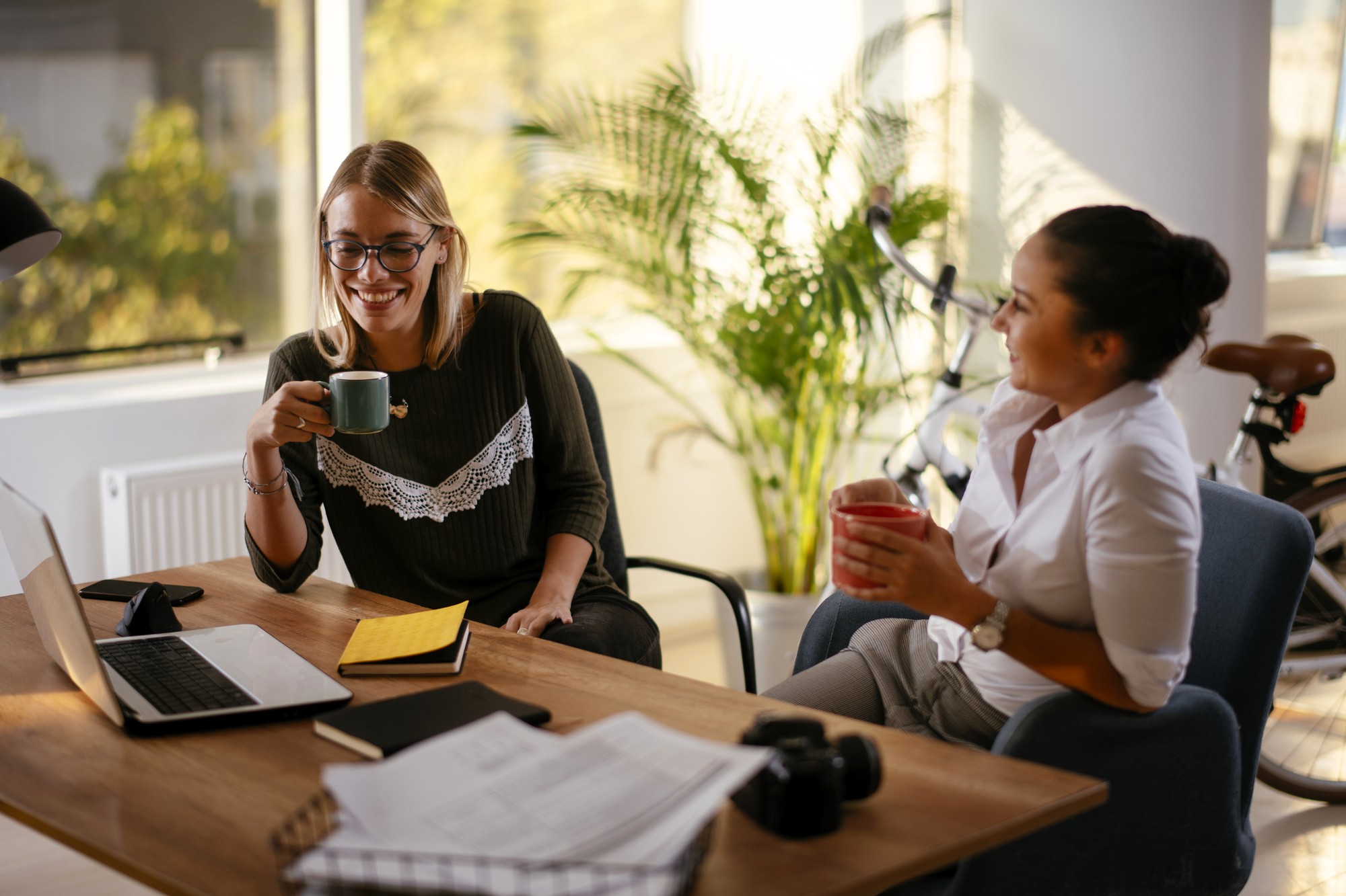 Two women sit at a desk in an office, smiling and chatting while holding mugs. Laptops, notebooks, and documents are on the desk, and a bicycle and plant are visible in the background.