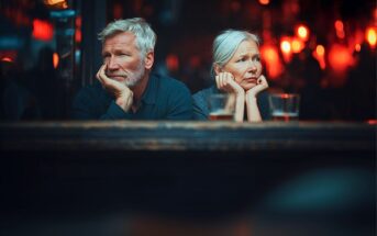 An older man and woman with gray hair sit side by side at a bar, both resting their faces in their hands and gazing thoughtfully ahead. The background is dimly lit with warm, blurry lights. Drinks sit on the table in front of them.