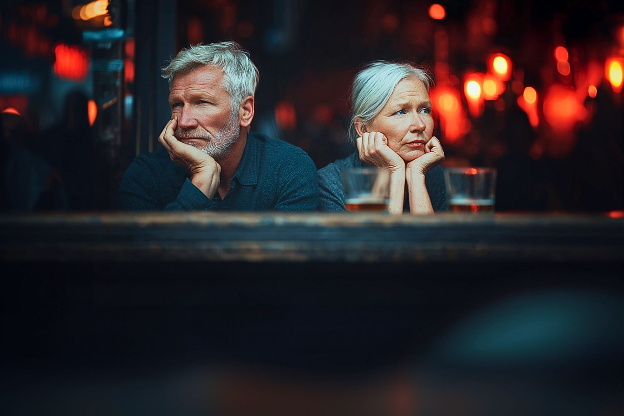 An older man and woman with gray hair sit side by side at a bar, both resting their faces in their hands and gazing thoughtfully ahead. The background is dimly lit with warm, blurry lights. Drinks sit on the table in front of them.
