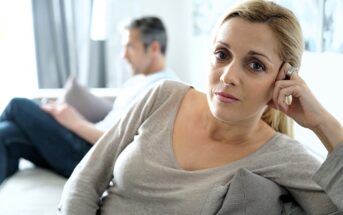 9 Reasons People Choose The Stability Of An Unhappy Marriage Instead Of Getting Divorced A woman sits on a couch looking concerned, resting her head on her hand. In the background, a man sits turned away, appearing distant and focused elsewhere. Both look serious and disconnected.