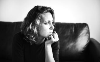 Black and white photo of a woman with short, wavy hair sitting on a couch, resting her chin on her hand, and gazing thoughtfully to the right. The background is plain and softly lit.