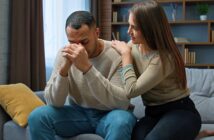 A woman sits beside a sad man on a couch, comforting him by gently touching his shoulder. The man has his head bowed and hands clasped in front of his face, while the woman looks at him with concern.