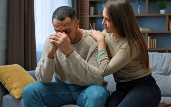 A woman sits beside a sad man on a couch, comforting him by gently touching his shoulder. The man has his head bowed and hands clasped in front of his face, while the woman looks at him with concern.