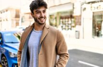 A young man with dark hair and a beard smiles while standing outdoors on a city street. He is wearing a light gray shirt and a tan coat. A blue car and blurred buildings are visible in the background.