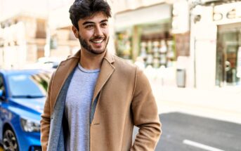 A young man with dark hair and a beard smiles while standing outdoors on a city street. He is wearing a light gray shirt and a tan coat. A blue car and blurred buildings are visible in the background.