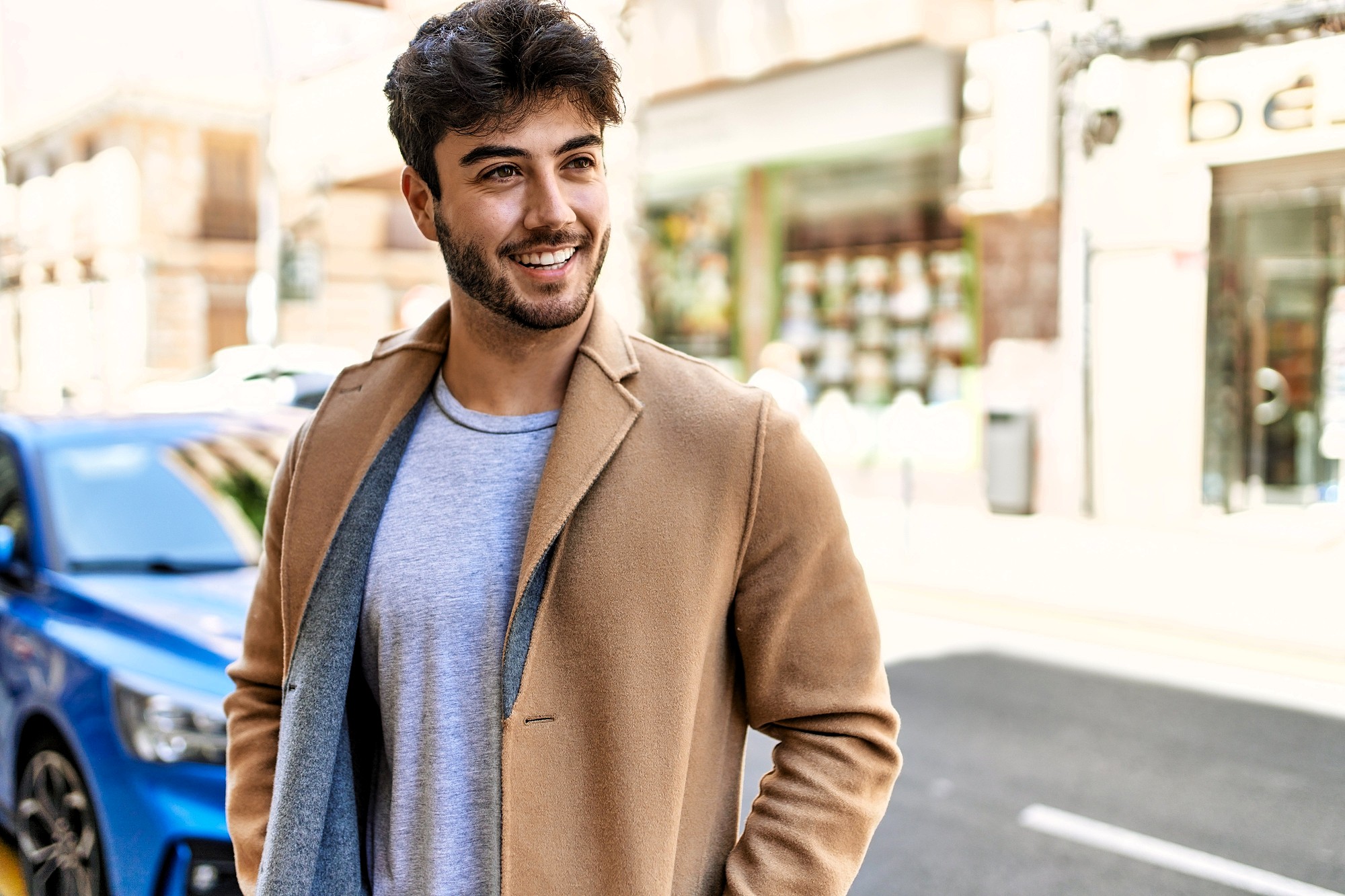 A young man with dark hair and a beard smiles while standing outdoors on a city street. He is wearing a light gray shirt and a tan coat. A blue car and blurred buildings are visible in the background.