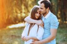 A young couple stands close together outdoors, with the man gently hugging the woman from behind. They both appear happy and content, surrounded by greenery and warm sunlight.