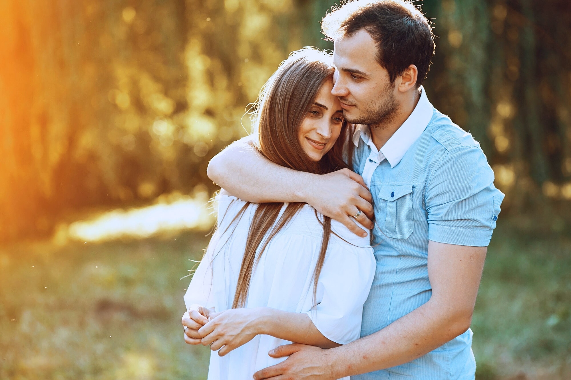 A young couple stands close together outdoors, with the man gently hugging the woman from behind. They both appear happy and content, surrounded by greenery and warm sunlight.