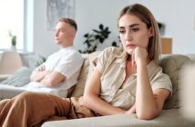 A woman sits on a couch looking sad and thoughtful, resting her chin on her hand, while a man sits in the background with his arms crossed, both appearing upset in a bright living room.