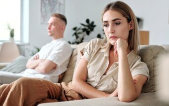 A woman sits on a couch looking sad and thoughtful, resting her chin on her hand, while a man sits in the background with his arms crossed, both appearing upset in a bright living room.