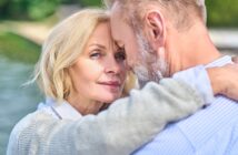 A close-up of an older couple embracing outdoors, with the woman looking at the camera and the man facing her, partially out of frame. They appear affectionate and content by the water.