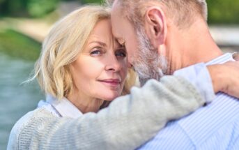 A close-up of an older couple embracing outdoors, with the woman looking at the camera and the man facing her, partially out of frame. They appear affectionate and content by the water.