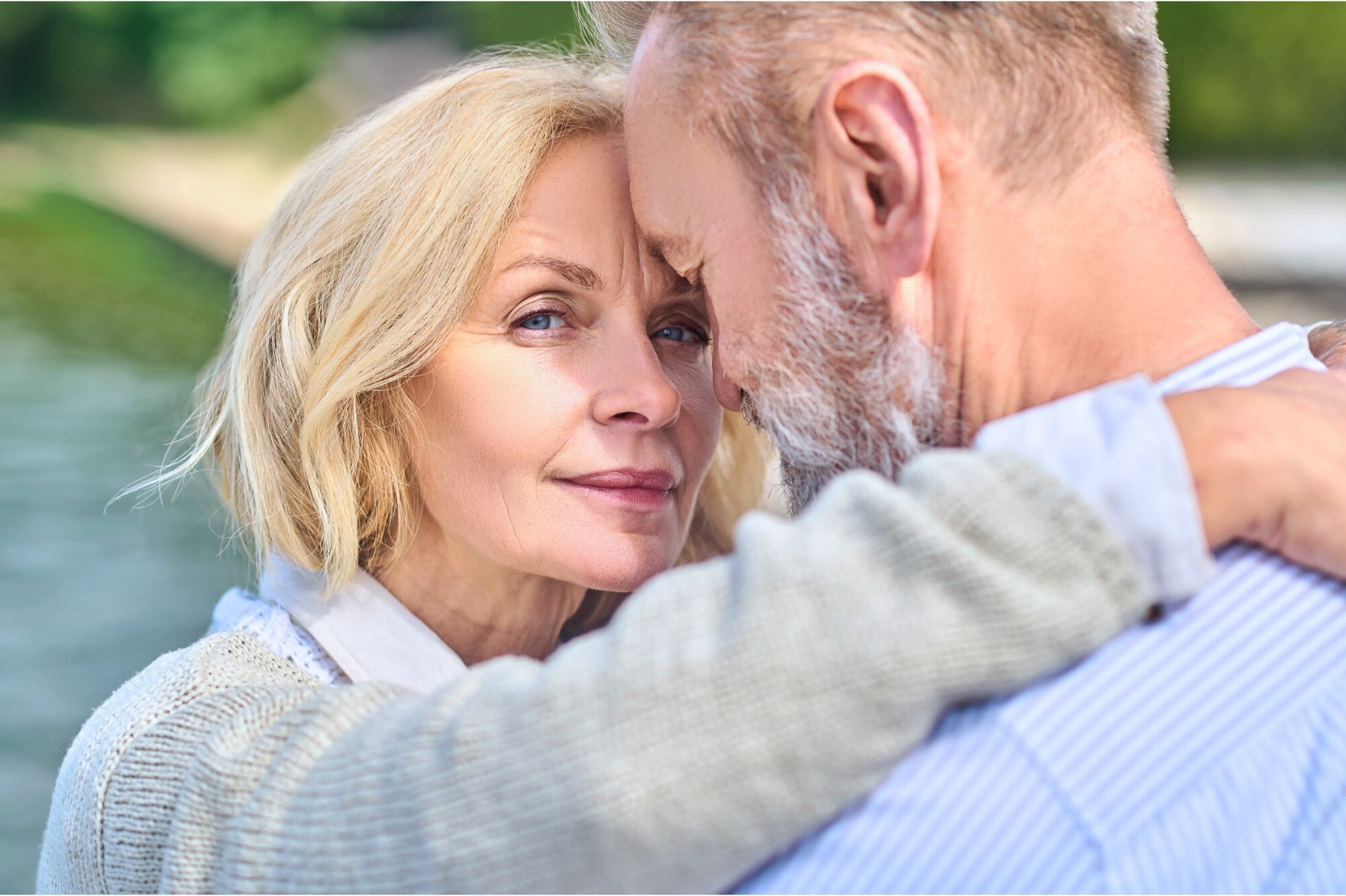 A close-up of an older couple embracing outdoors, with the woman looking at the camera and the man facing her, partially out of frame. They appear affectionate and content by the water.