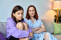 A teenage girl in a purple hoodie sits on a couch, hugging her knees and looking upset. Beside her, a woman with red hair in a striped dress sits with folded arms, watching her with a concerned expression.