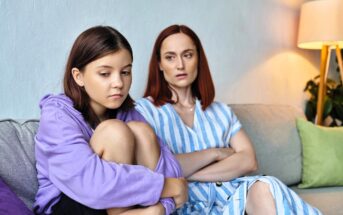 A teenage girl in a purple hoodie sits on a couch, hugging her knees and looking upset. Beside her, a woman with red hair in a striped dress sits with folded arms, watching her with a concerned expression.