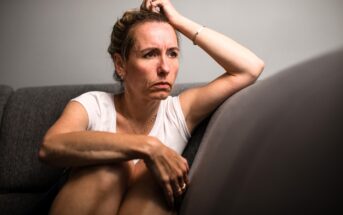 A woman sits on a gray couch with a serious, pensive expression, resting her arm on her head and hugging her knees, looking off to the side in a contemplative mood.
