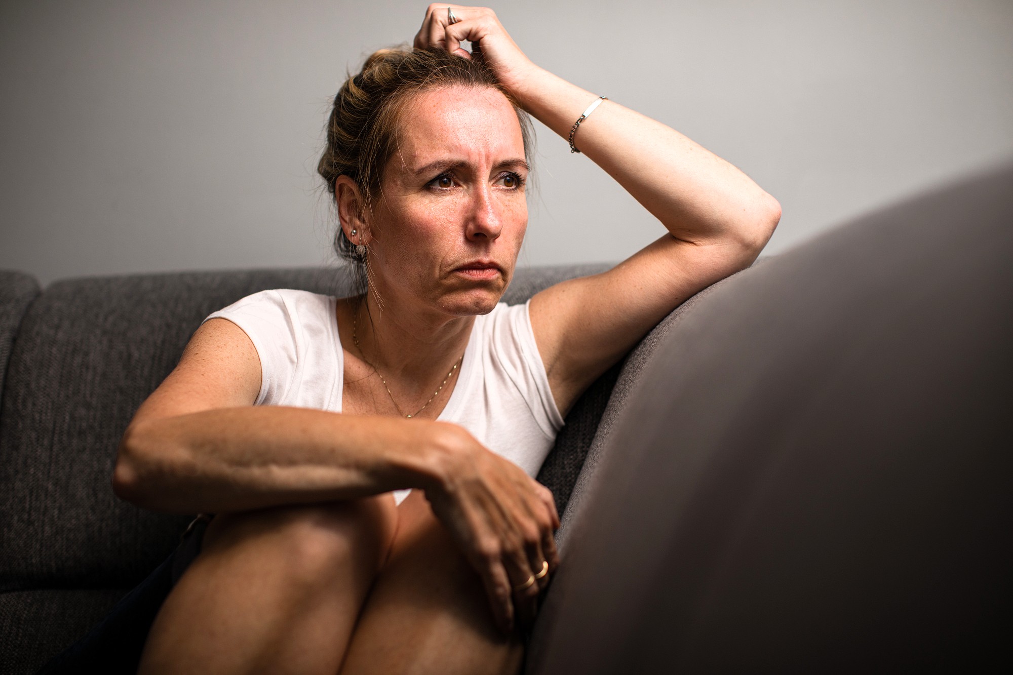 A woman sits on a gray couch with a serious, pensive expression, resting her arm on her head and hugging her knees, looking off to the side in a contemplative mood.