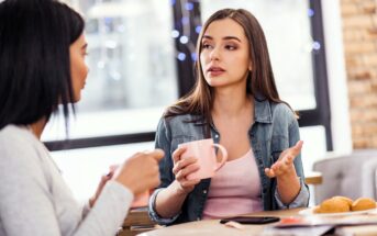 Two women sit at a table indoors, holding mugs and having a serious conversation. One gestures with her hand while speaking. There are pastries and a phone on the table, and soft lights in the background.