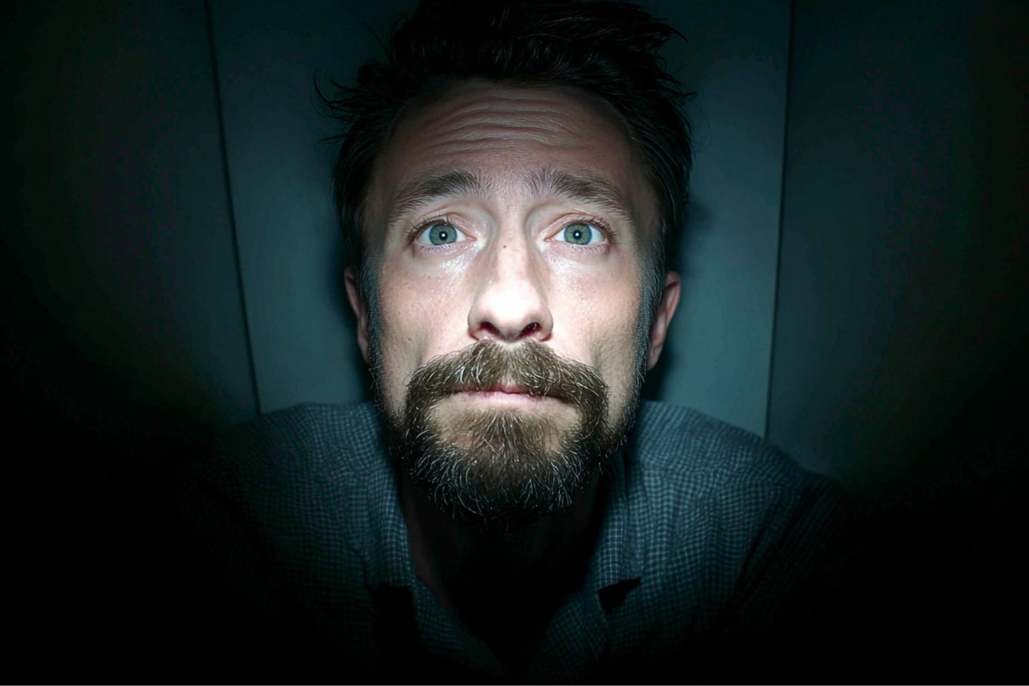 A man with light eyes and a beard looks up toward a light source in a dimly lit room, his expression appearing worried or anxious. Shadows surround his face, highlighting his features.