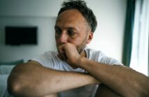 A man in a white t-shirt sits indoors with his arms resting on his knees, gazing thoughtfully to the side with a serious expression. The background is softly lit with a TV and a window visible.