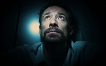 A man with a beard sits in darkness, looking up toward a bright light above him. The light casts shadows on his face, highlighting his serious and contemplative expression.