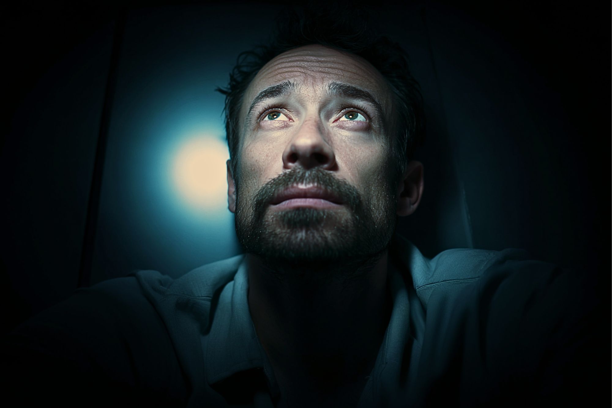 A man with a beard sits in darkness, looking up toward a bright light above him. The light casts shadows on his face, highlighting his serious and contemplative expression.