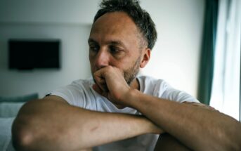 A man in a white t-shirt sits indoors with his arms resting on his knees, gazing thoughtfully to the side with a serious expression. The background is softly lit with a TV and a window visible.