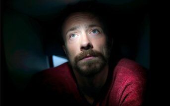 A man with a beard and mustache, wearing a red sweater, looks up into a beam of light in a dark room, with his face partially illuminated and an expression of contemplation or concern.